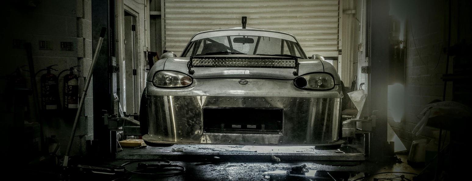 A stripped-back, bare-metal Mazda MX5 race car in a workshop.